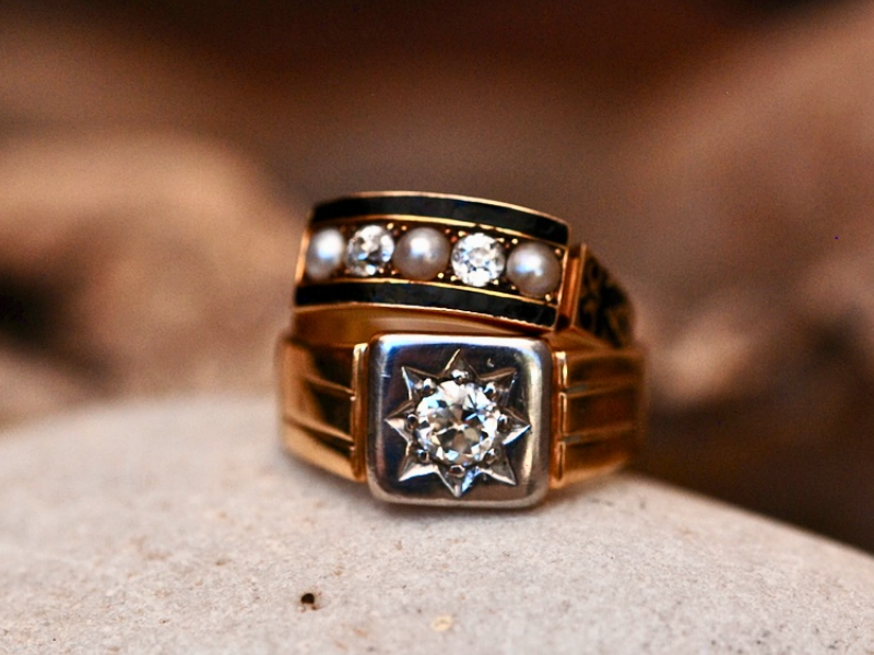 A photograph of an Edwardian 18ct yellow gold diamond signet ring in on a pebble with another Edwardian Onyx, gold, diamond and pearl ring stacked on top.