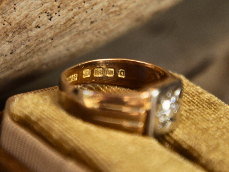A photograph of an Edwardian 18ct yellow gold diamond signet ring on lying on its side to view the stamps on a gold velvet ring box.