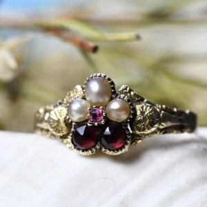 A picture of a gold Georgian Daisy Ring with two garnet petals and three pearl petals set into an engraved base of swirls surrounding a link pink garnet.